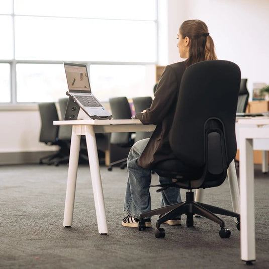 A woman sits in a black office chair at a white desk, viewed from behind as she works on a laptop in a bright, modern office space.