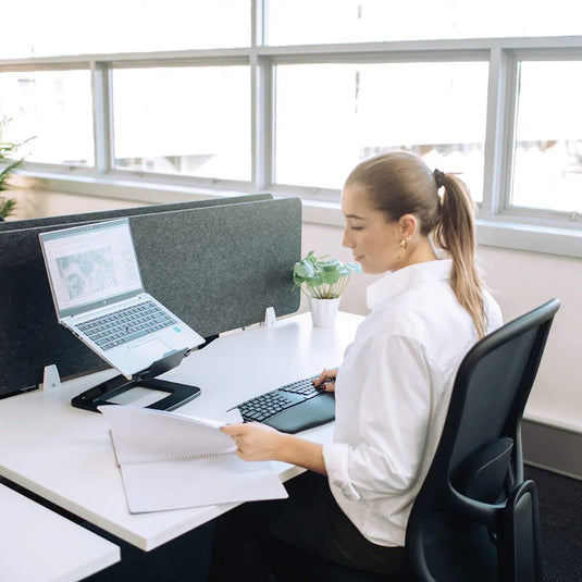 A woman in a white blouse sits at a modern office desk, working on a laptop and holding papers near large windows.