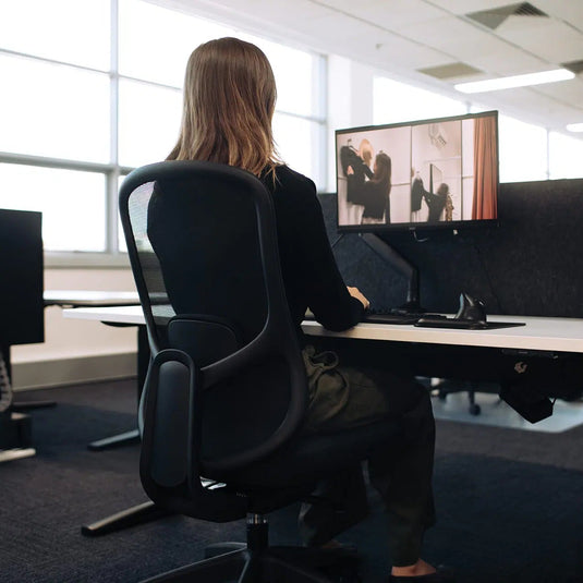 A woman sits in a black ergonomic office chair at a standing desk, viewed from behind as she looks at a computer monitor in a bright, modern office space.