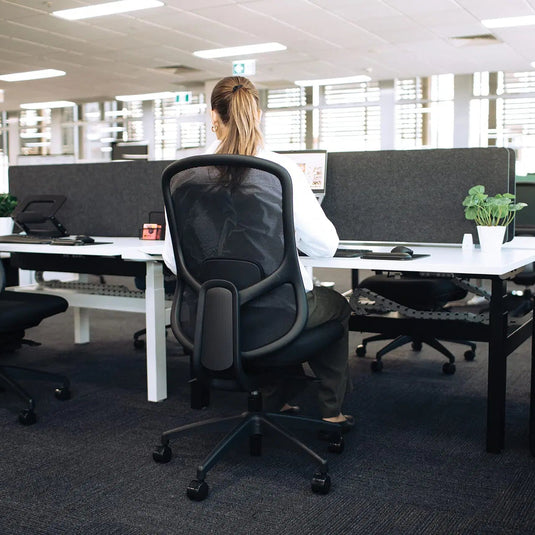 A woman sits at a desk in a modern office, viewed from behind as she works at a computer. The scene features black mesh office chairs, white desks, and gray cubicle dividers.