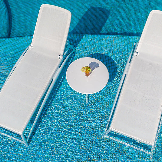 Two white lounge chairs and a small round table with a yellow object in the center, all on a pool deck.