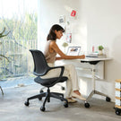 Person working at a desk with a chair in a bright room with large windows.