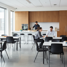 Modern kitchen and dining area with people interacting, large windows, and wooden cabinets.
