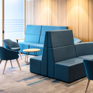 Modern office lounge seating area featuring a large modular blue sofa, a matching armchair, a smaller accent chair, and a round wooden side table on a patterned carpet in front of a wood-paneled wall.