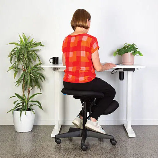 Load image into Gallery viewer, Person sitting on a black office chair at a white desk with plants around.
