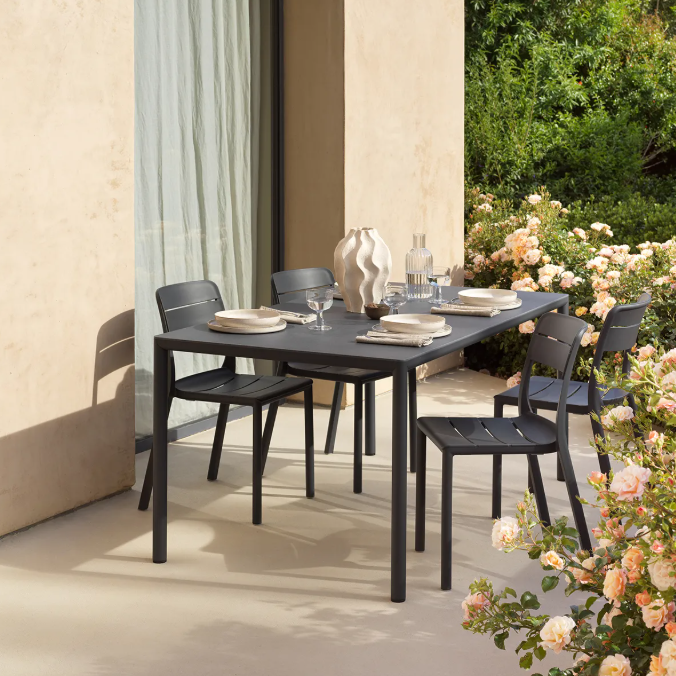 Load image into Gallery viewer, Outdoor dining setup with a black table and chairs on a patio with flowers and a building in the background.
