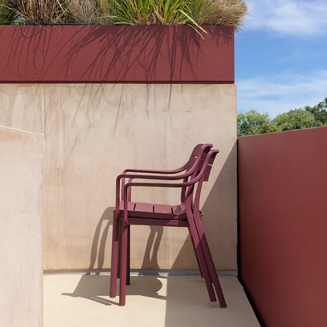 Load image into Gallery viewer, Maroon chairs against a beige wall with a planter of grasses on a sunny day.
